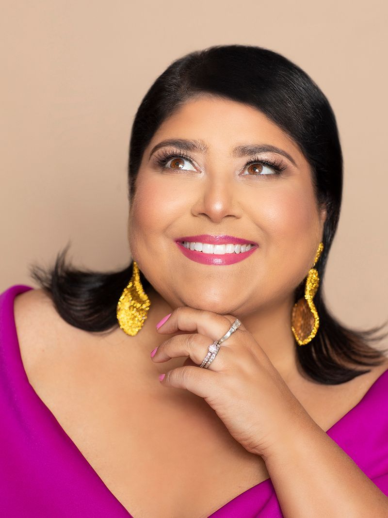 Smiling person in a magenta top with gold earrings, resting chin on hand against a beige background