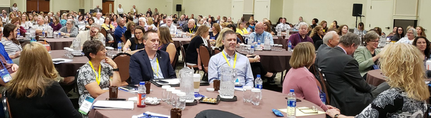 A large audience seated at round tables in a conference hall during a presentation or meeting.
