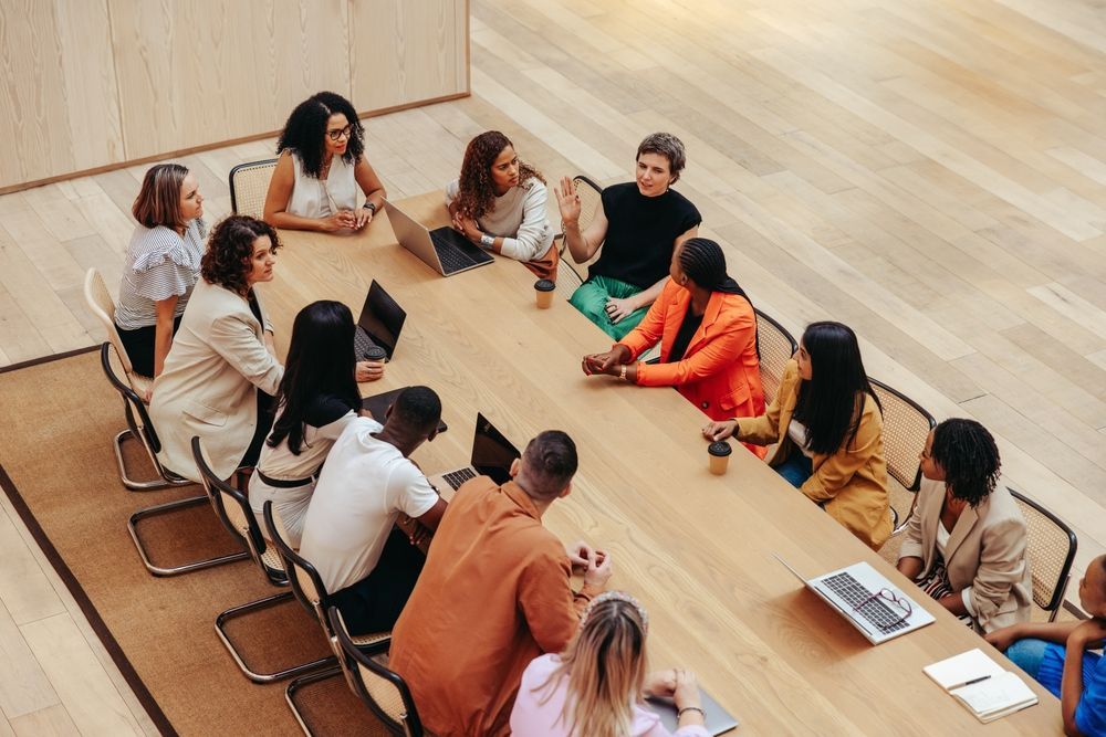 A diverse group of people sits around a large wooden table in a brightly lit office for a collaborative meeting.