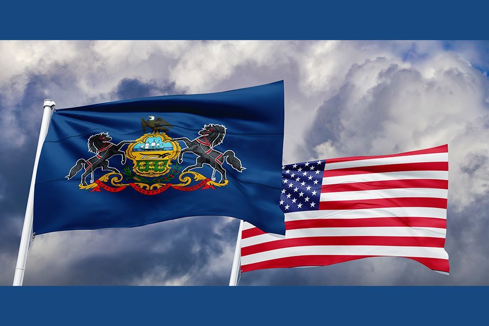 The state flag of Pennsylvania flying beside the American flag against a cloudy sky.
