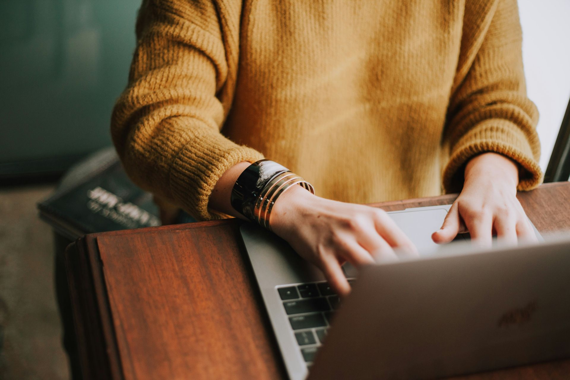 A person wearing a mustard yellow sweater types on a laptop at a wooden desk, wearing a stacked bracelet on their wrist.