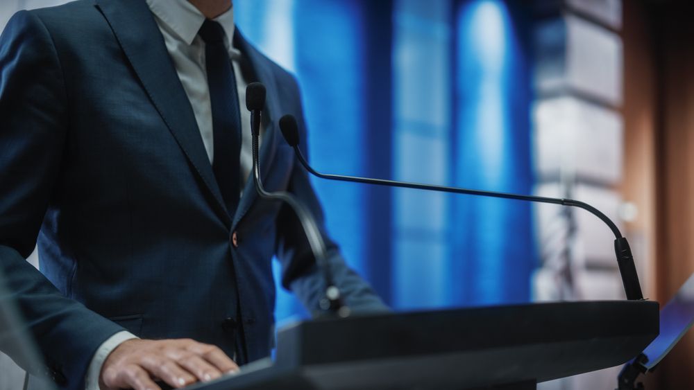A person in a blue suit stands at a podium with microphones in front of a blue-lit backdrop.