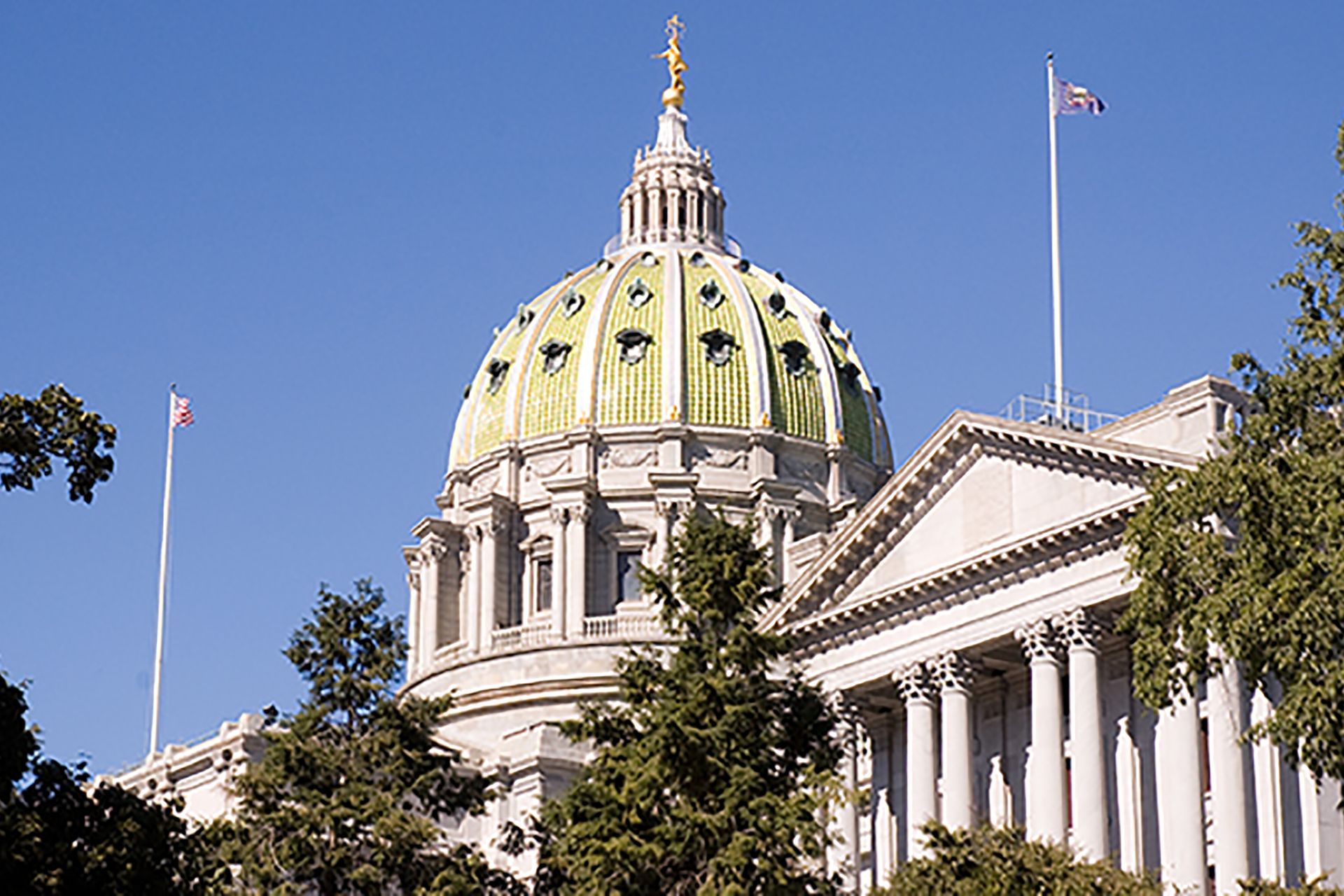 The domed Pennsylvania State Capitol building with green copper accents, seen behind trees against a clear blue sky.