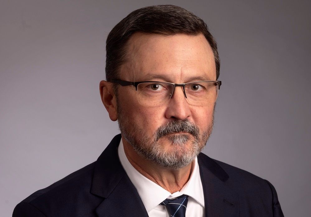Headshot of a person with short brown hair and glasses, wearing a dark suit, white shirt, and blue tie against a grey.
