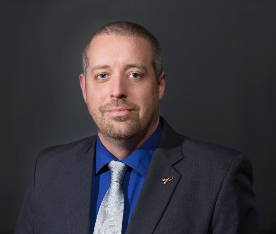 A professional headshot of a man wearing a dark grey suit, blue dress shirt, and patterned tie against a grey background.