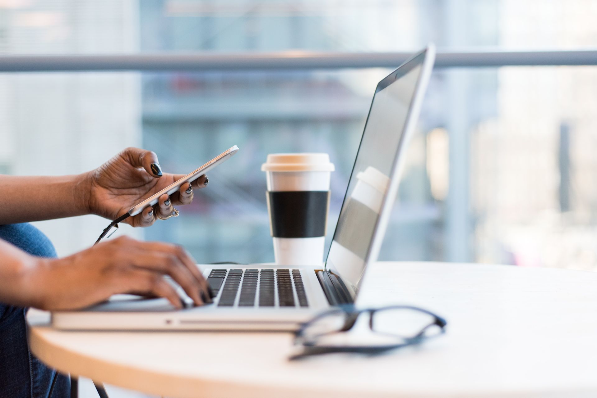 Black woman at laptop with phone in hand, coffee, and glasses on table