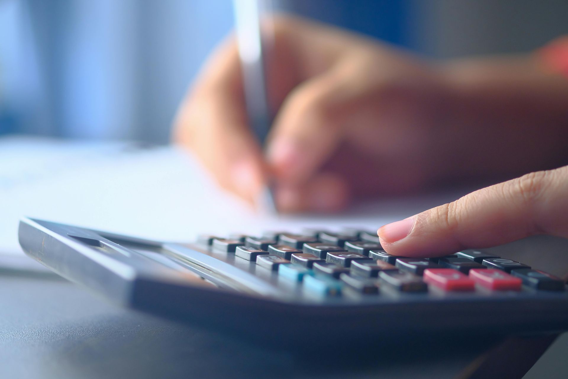 A person’s hand pressing buttons on a calculator while they hold a pen to write on paper.