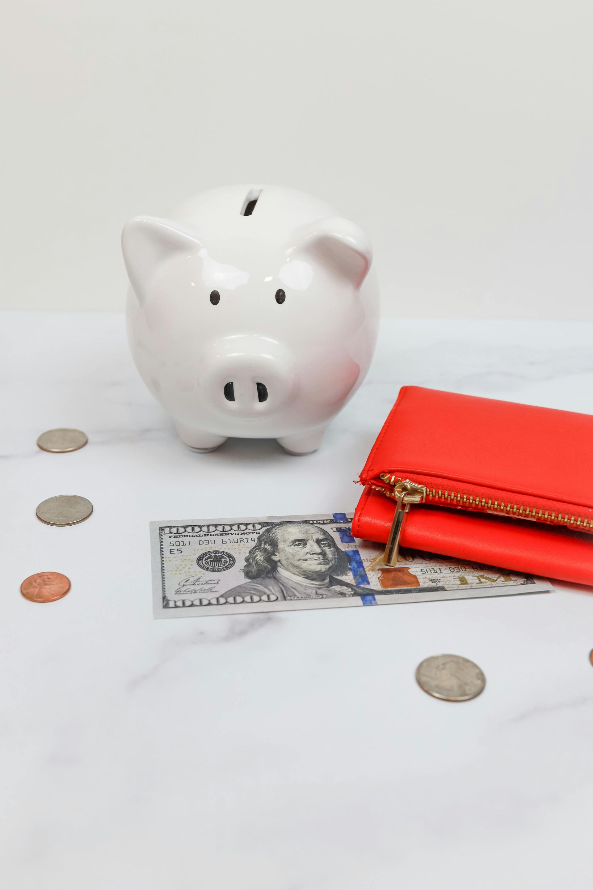 A white piggy bank, a hundred-dollar bill, and a red wallet on a marble surface with scattered coins.