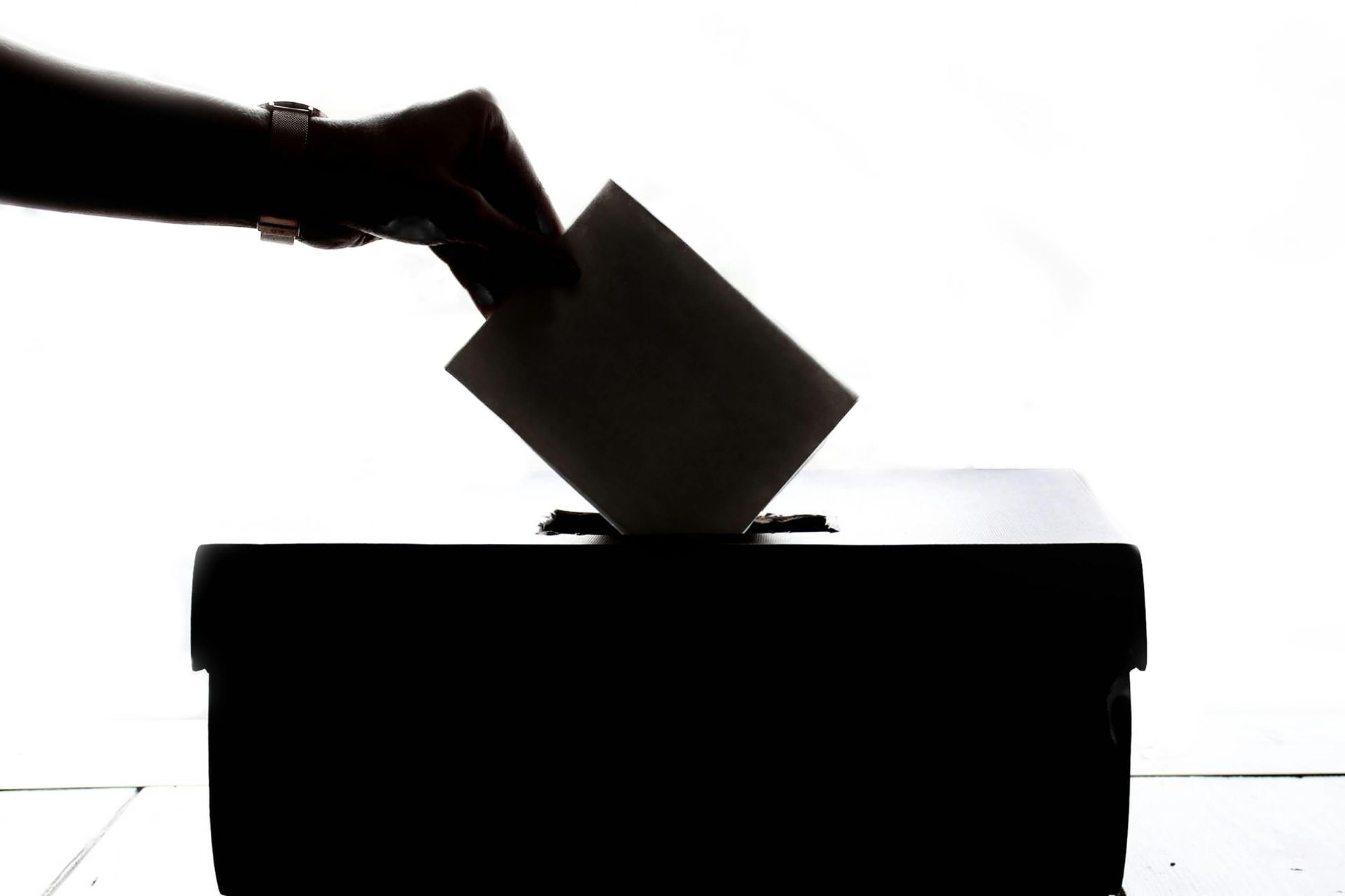 A silhouette of a hand placing a ballot paper into a ballot box against a bright white background.