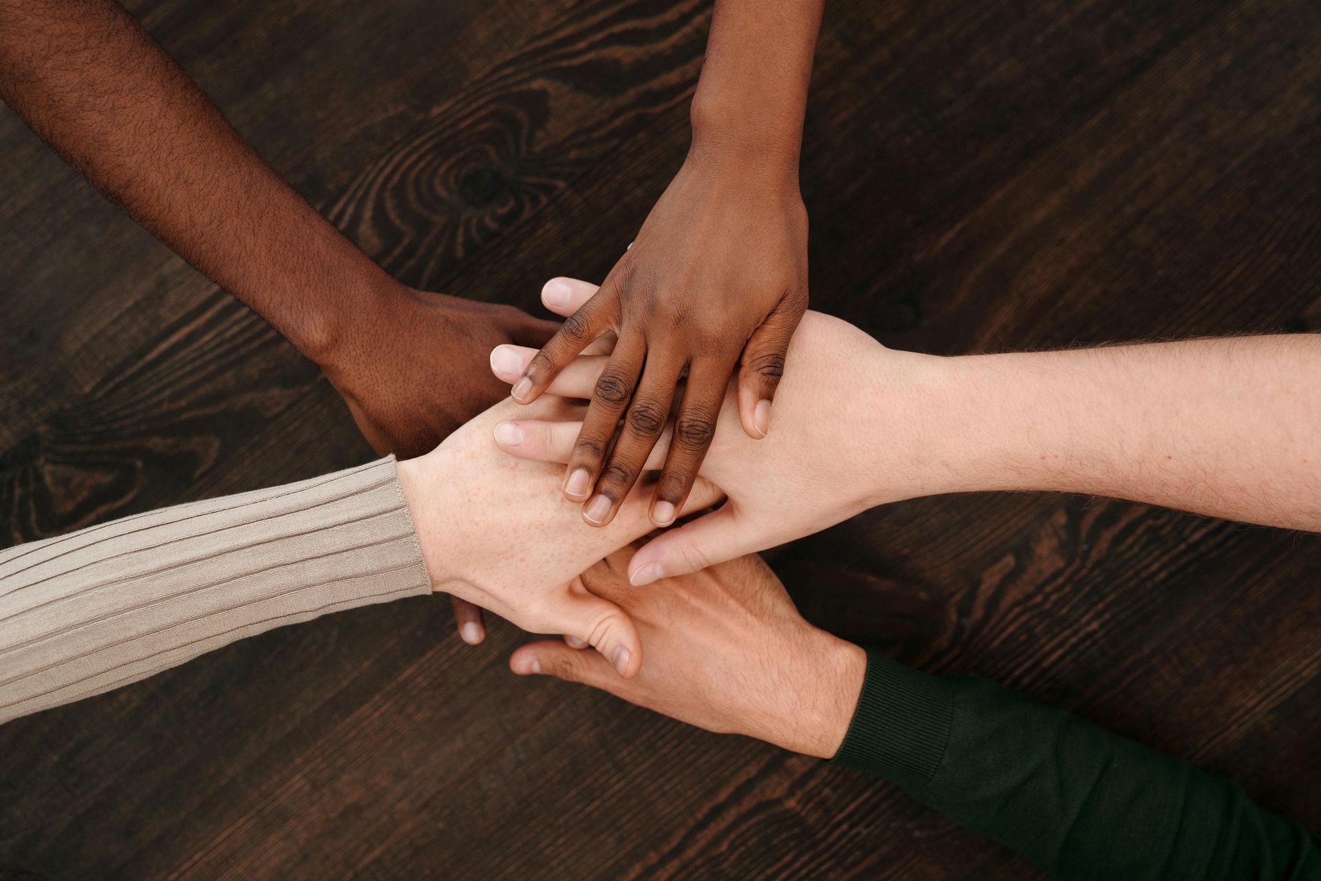 Four people place their hands together in the center against a dark wooden background, symbolizing unity and cooperation.