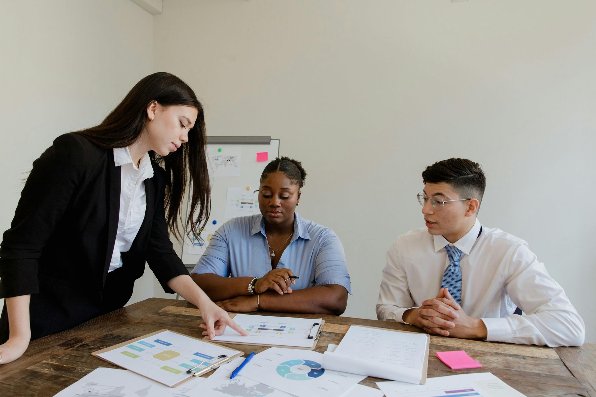 Three people sit at a wooden table in an office, reviewing documents and discussing work materials.