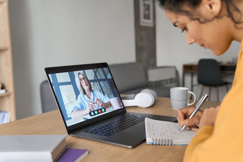 A person wearing a yellow top takes notes while watching a video call with a colleague on a laptop at a wooden desk.