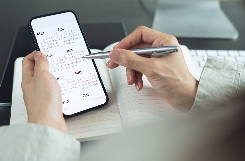 Hands holding a smartphone displaying a digital calendar while using a pen to write on an open notebook.