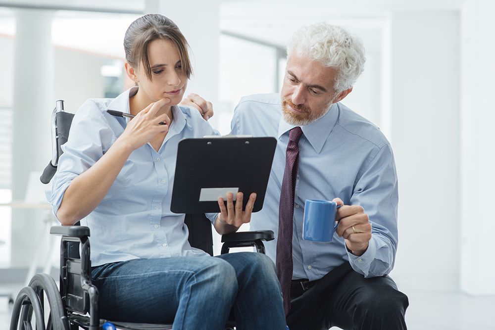 Woman in wheelchair looking at work tablet with manager kneeling next to her