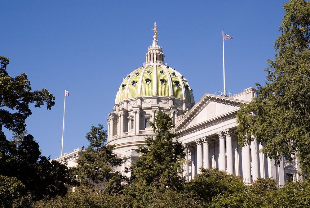 The Pennsylvania State Capitol building features a large green dome and white columns, framed by green trees against a sky.
