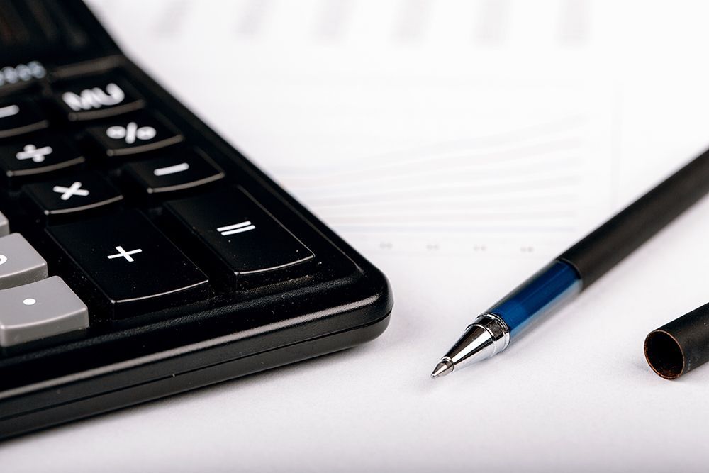 A black calculator and a blue pen resting on a white surface with blurred financial charts in the background.