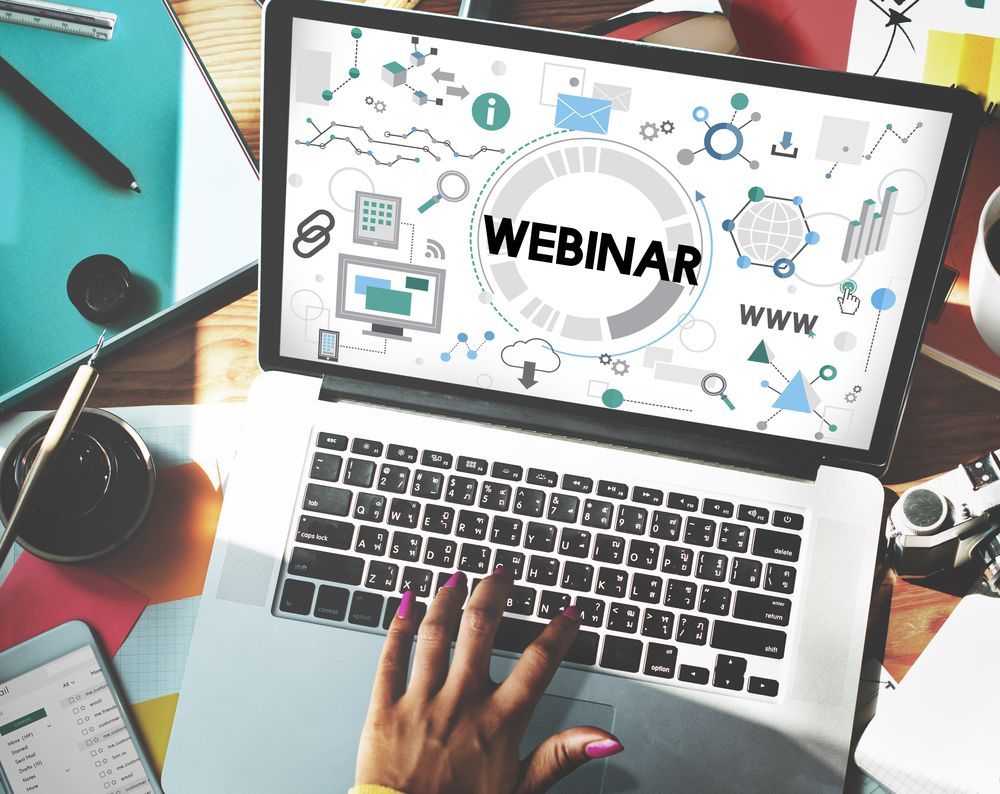 Laptop showing a webinar slide, with hands typing at a desk surrounded by notebooks and office supplies