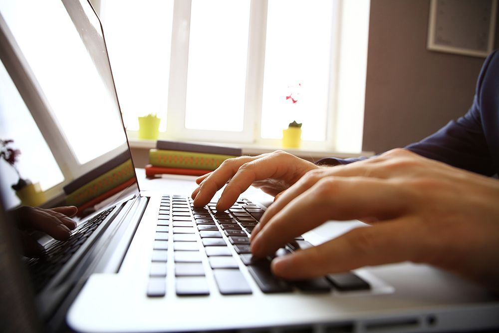 Close-up of hands typing on a laptop keyboard in a bright office space with books on a desk near a window.