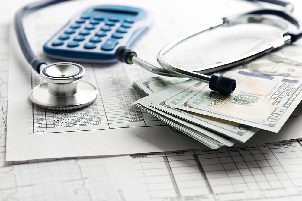 A stethoscope and a blue calculator resting on medical charts next to a stack of US one-hundred-dollar bills.