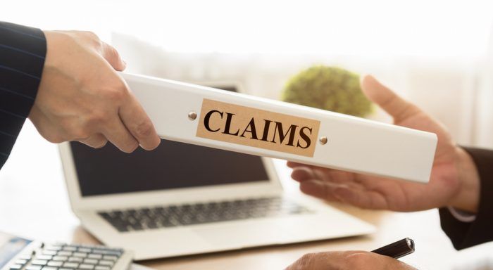 Two hands exchanging a folder labeled “CLAIMS” over a laptop desk.
