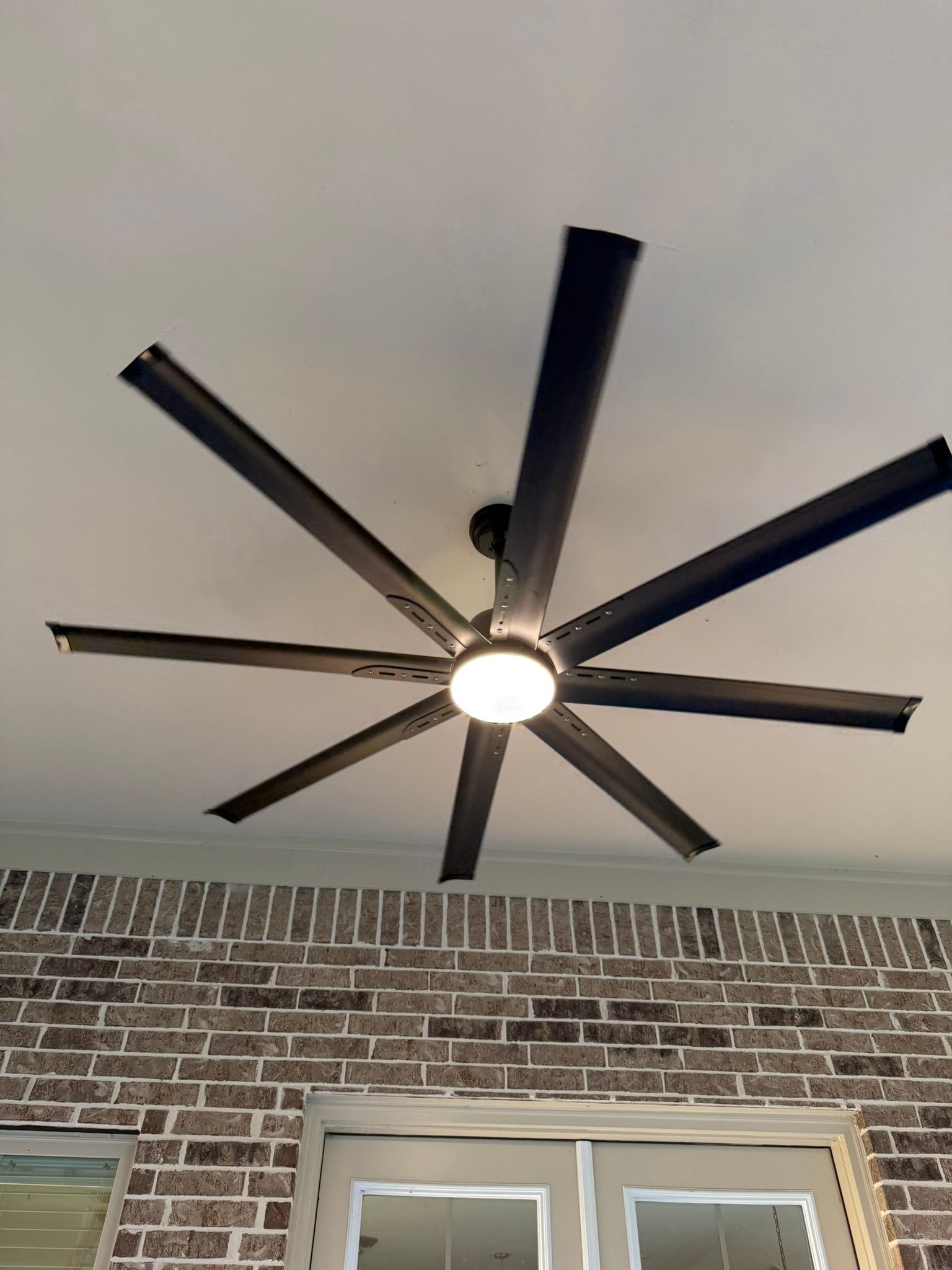 Ceiling fan with black blades, lit center, mounted on white ceiling, over brick wall, near door.