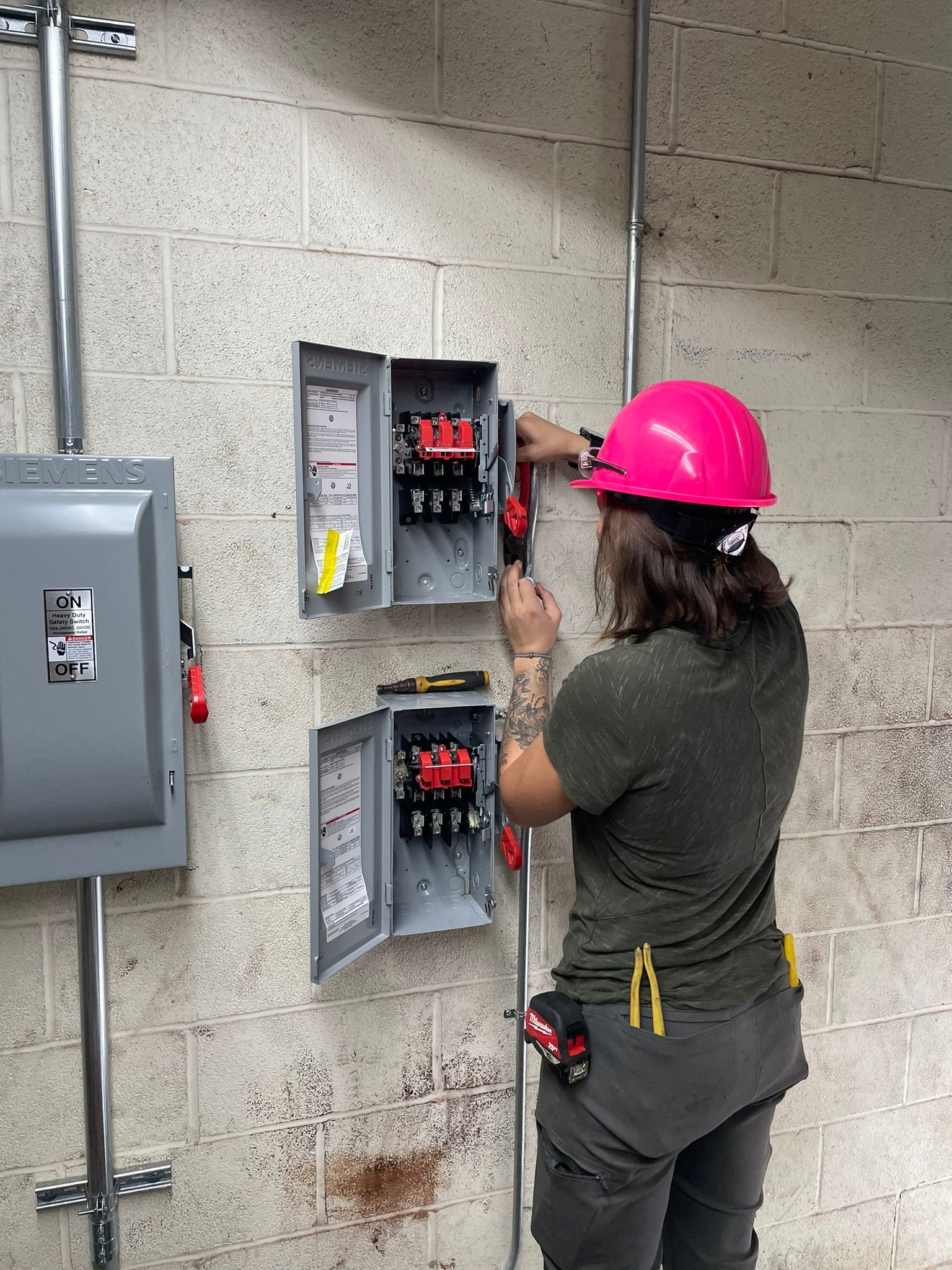 Woman in pink hard hat works on electrical panel mounted on a brick wall.
