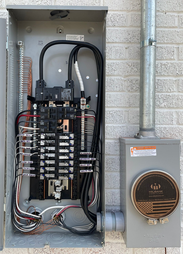 Gray electrical panel on a brick wall with wires and a meter to the right.