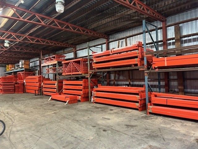 Une zone de stockage d'entrepôt avec des étagères industrielles métalliques supportant des piles de poutres de soutien horizontales orange.