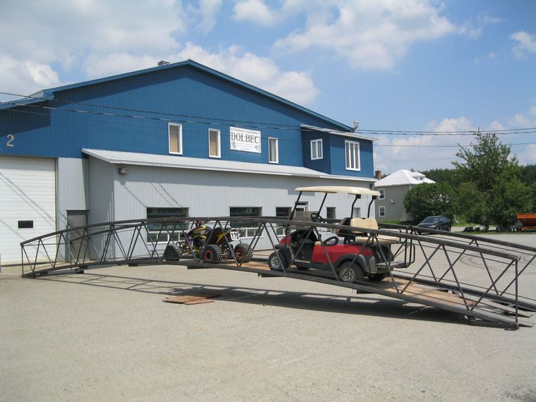 Une voiturette de golf rouge et un petit quad sont posés sur une structure de pont métallique arquée préfabriquée, garée sur un terrain gravillonné près d'un bâtiment bleu.