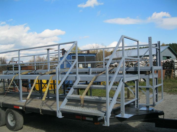 Plateforme de travail industrielle en métal gris, avec escaliers et garde-corps de sécurité, montée sur une remorque à plateau à l'extérieur.