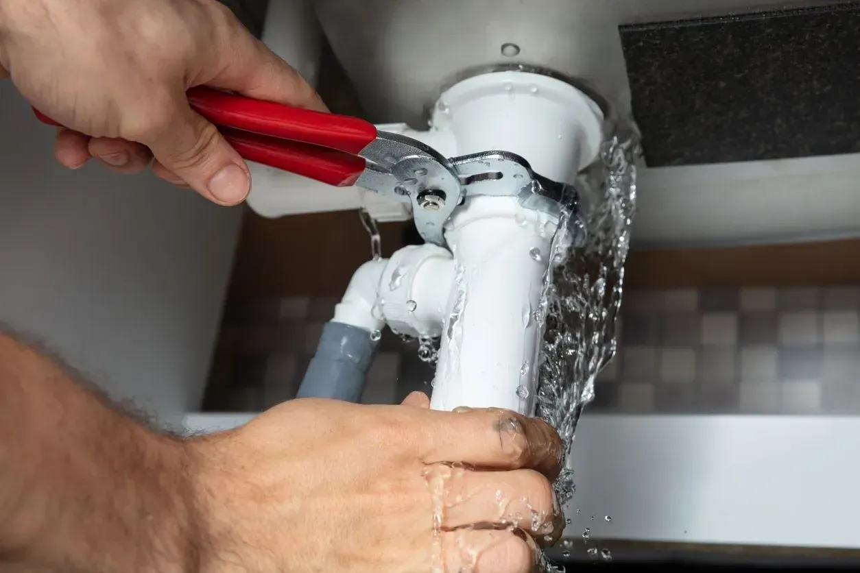 Hands using red pliers to repair a leaking white pipe under a sink