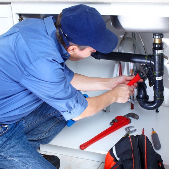 A professional in a blue uniform kneels under a sink, using a red pipe wrench to repair plumbing beneath the basin.