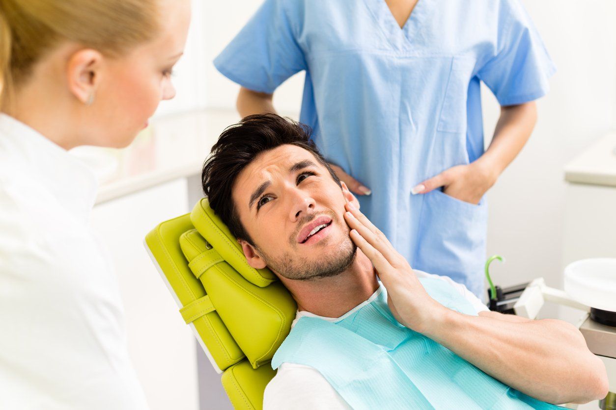 man-in-dental-chair-holds-painful-jaw-while-dentist-talks