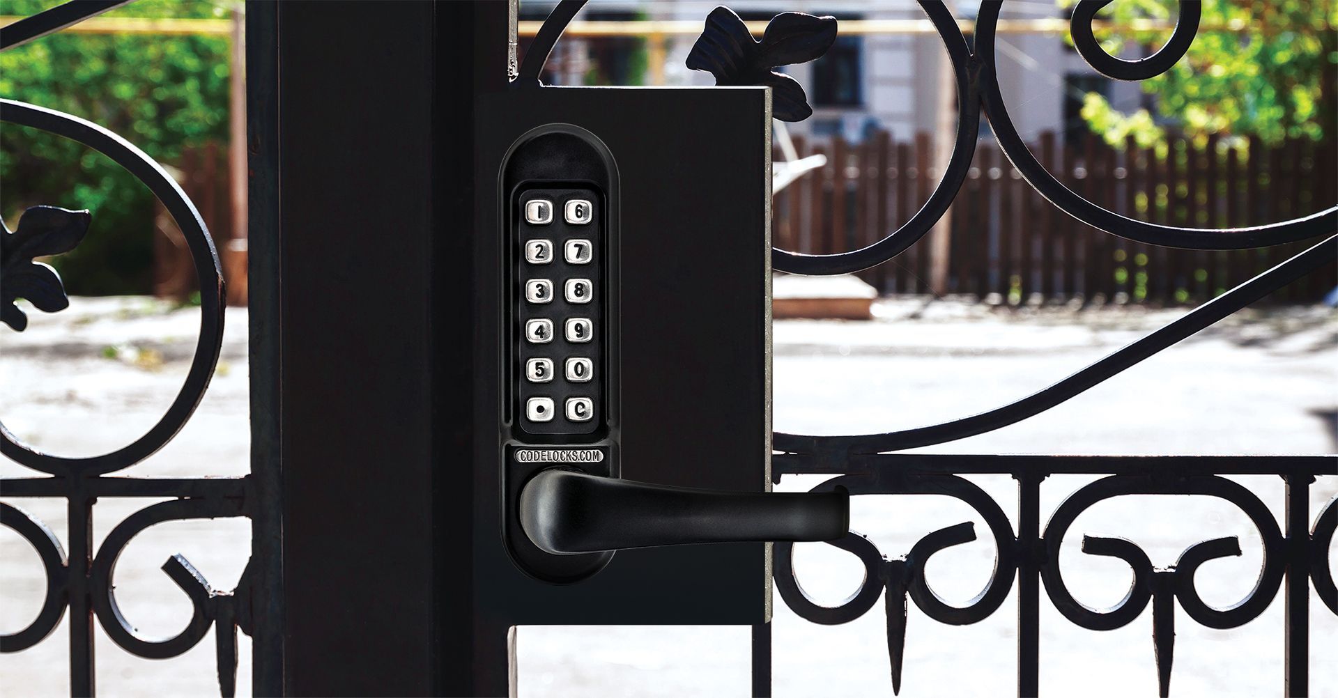 Black keypad lock and handle on a black gate, with decorative scrollwork, set outdoors.