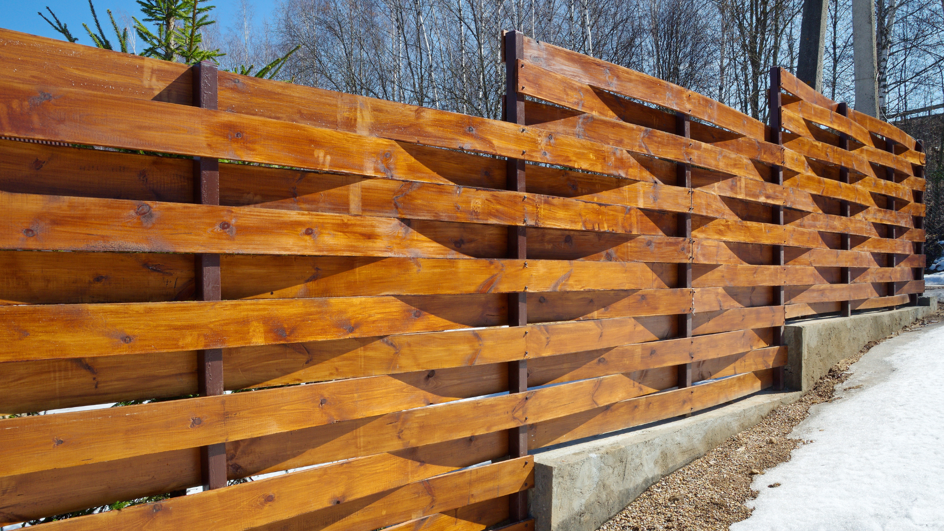 Wooden fence with a weaving pattern, stained a warm brown, along a snowy path.