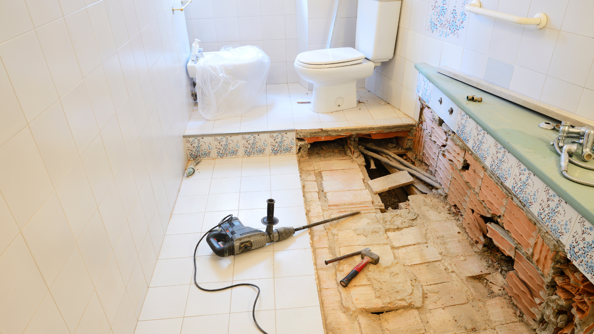Bathroom renovation in progress; exposed floor, brick wall, toilet, drill on floor, debris, and tools.