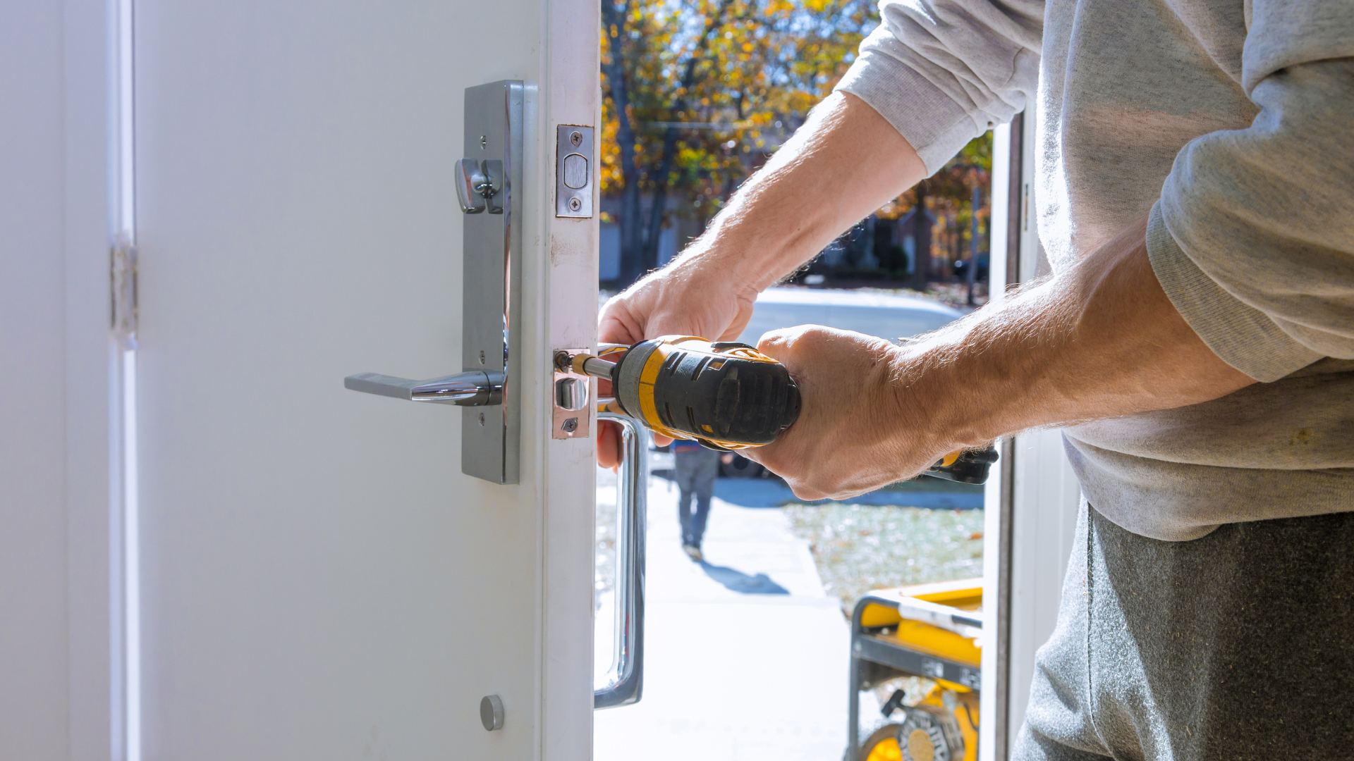 Person using a power drill to install a new lock on a white door. Outdoors, sunny setting.