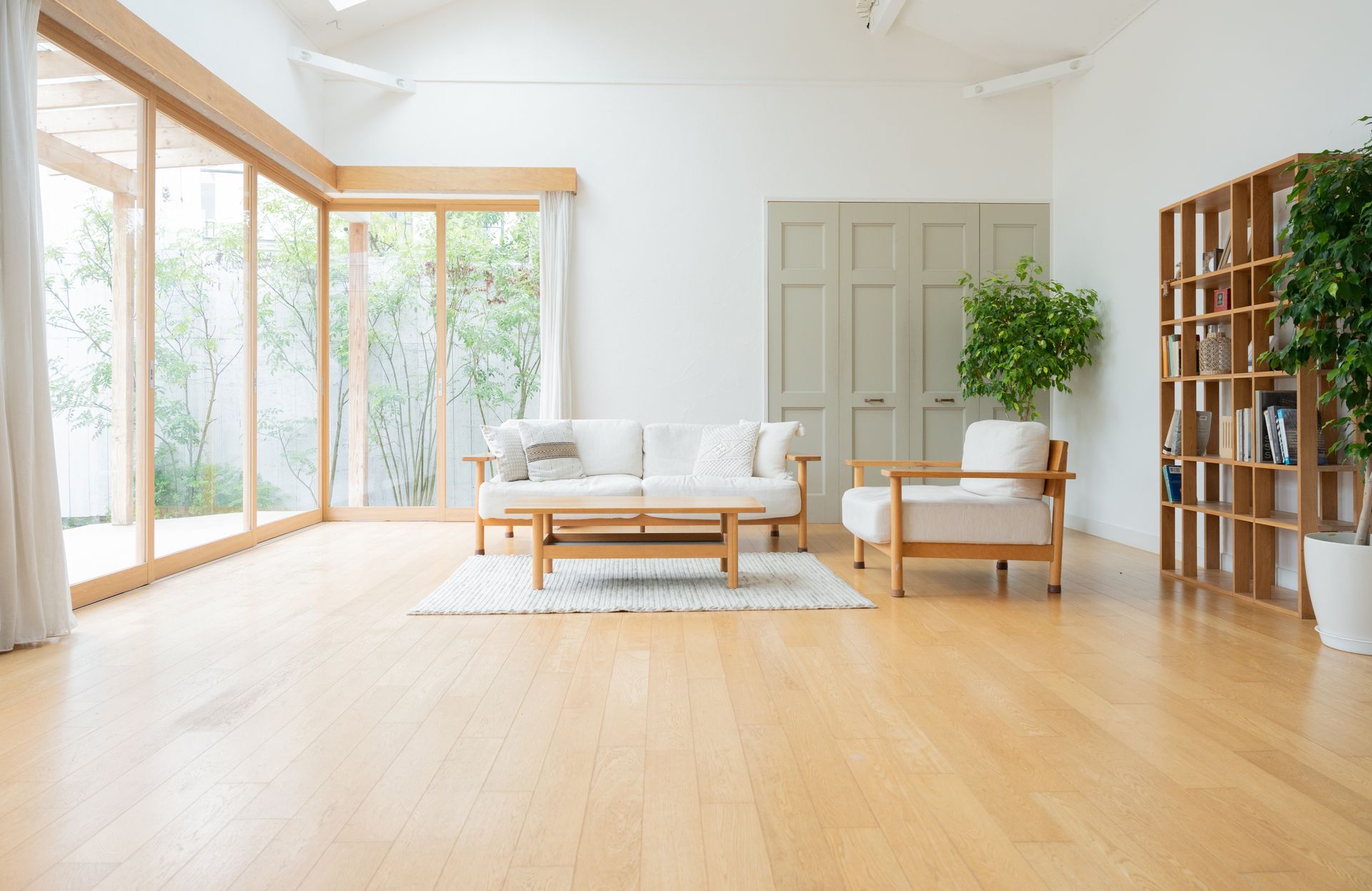 Bright living room with wood furniture, large windows, white walls, and plants.