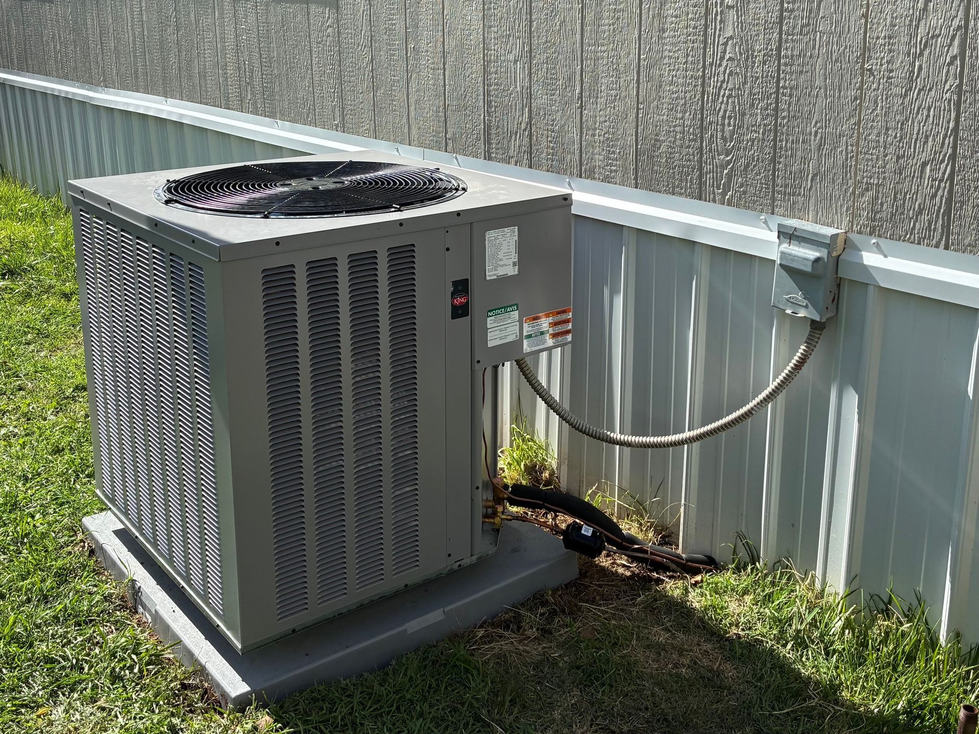 An air conditioning unit sits on a concrete pad next to a fence in a grassy yard.