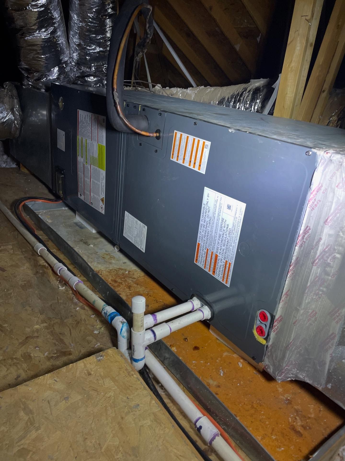 HVAC unit in an attic with PVC pipes for drainage. Gray metal unit, insulation, wooden floor.