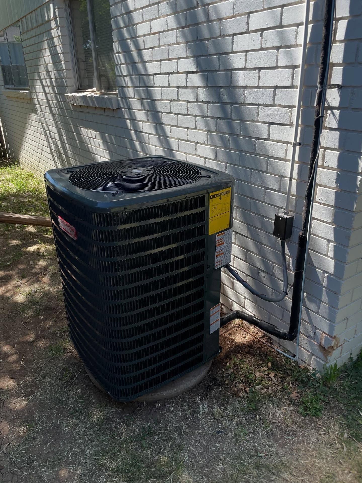 An air conditioning unit next to a light-colored brick building. Black metal, yellow label, and a dark pipe.