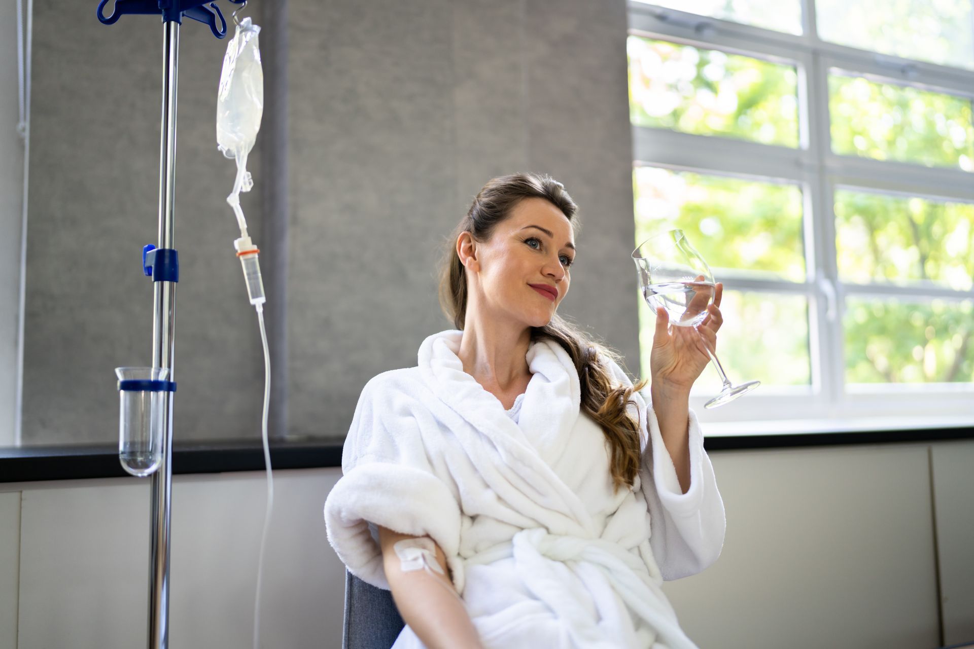 a woman in a bathrobe is sitting in a hospital chair holding a glass of water .