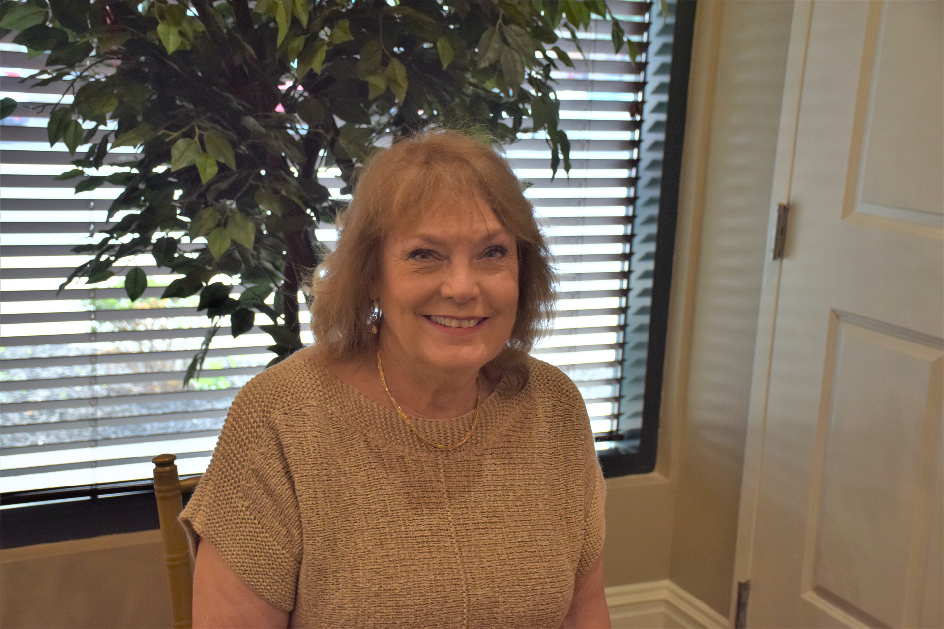A woman is sitting in front of a window with blinds and smiling.