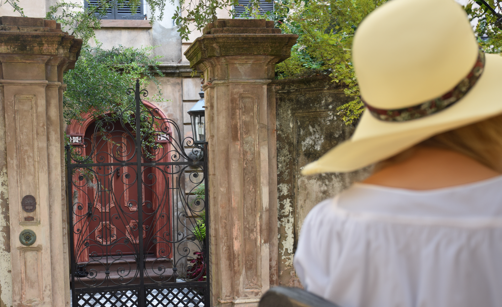 Woman in a sunhat looks toward an ornate gate and doorway with aged columns in an outdoor setting.