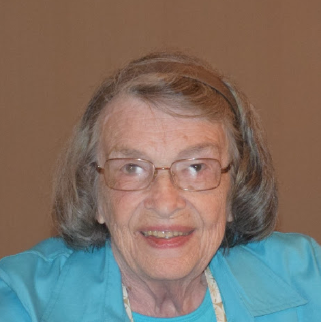 An elderly woman wearing glasses and a blue shirt smiles for the camera