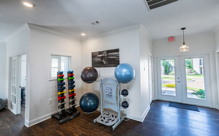 Gym interior with weights, exercise balls, and exit doors. Dark wood floors, white walls, bright lighting.