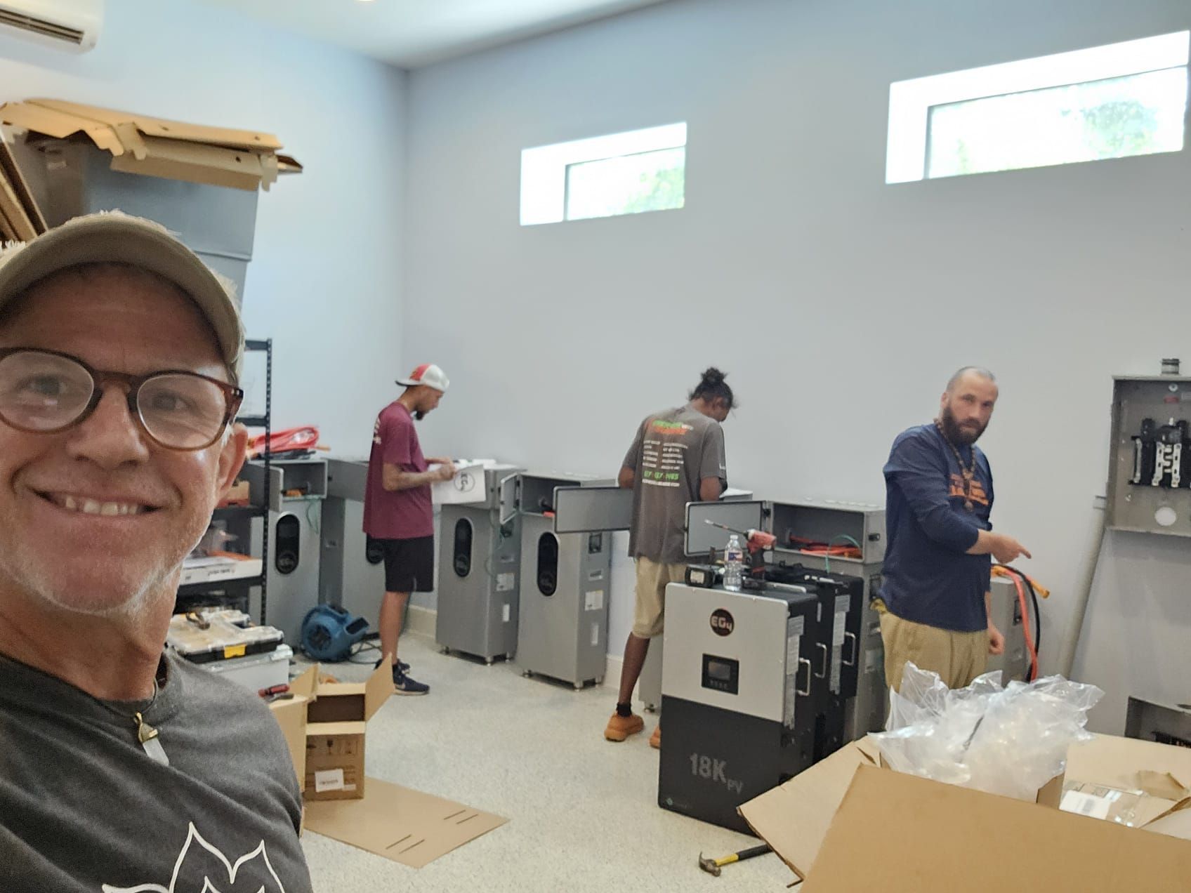 A man is taking a selfie in a room with other men working on a machine.