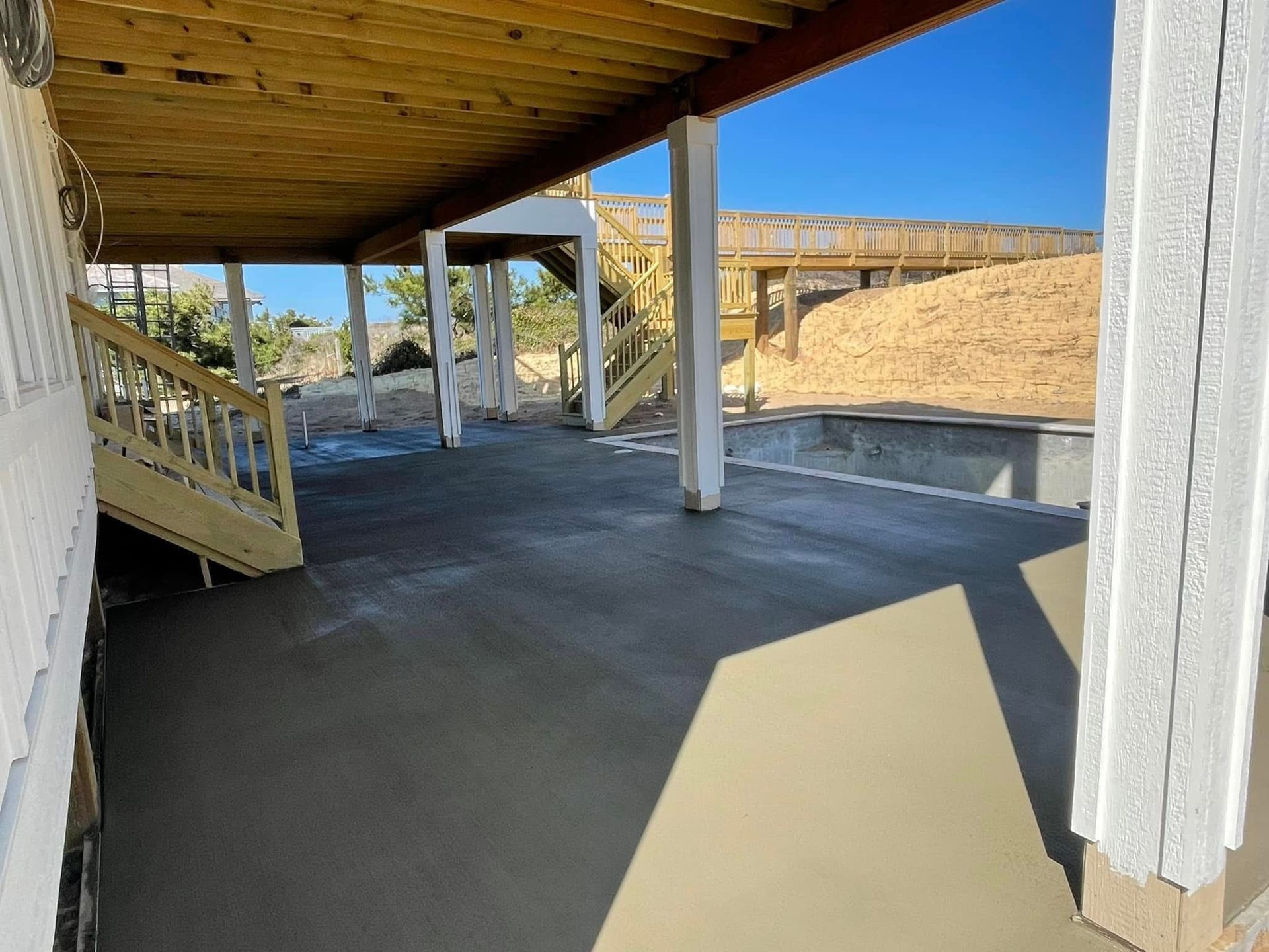 Underneath a deck, a concrete patio with stairs, support beams, and a pile of wood. Sunny day.