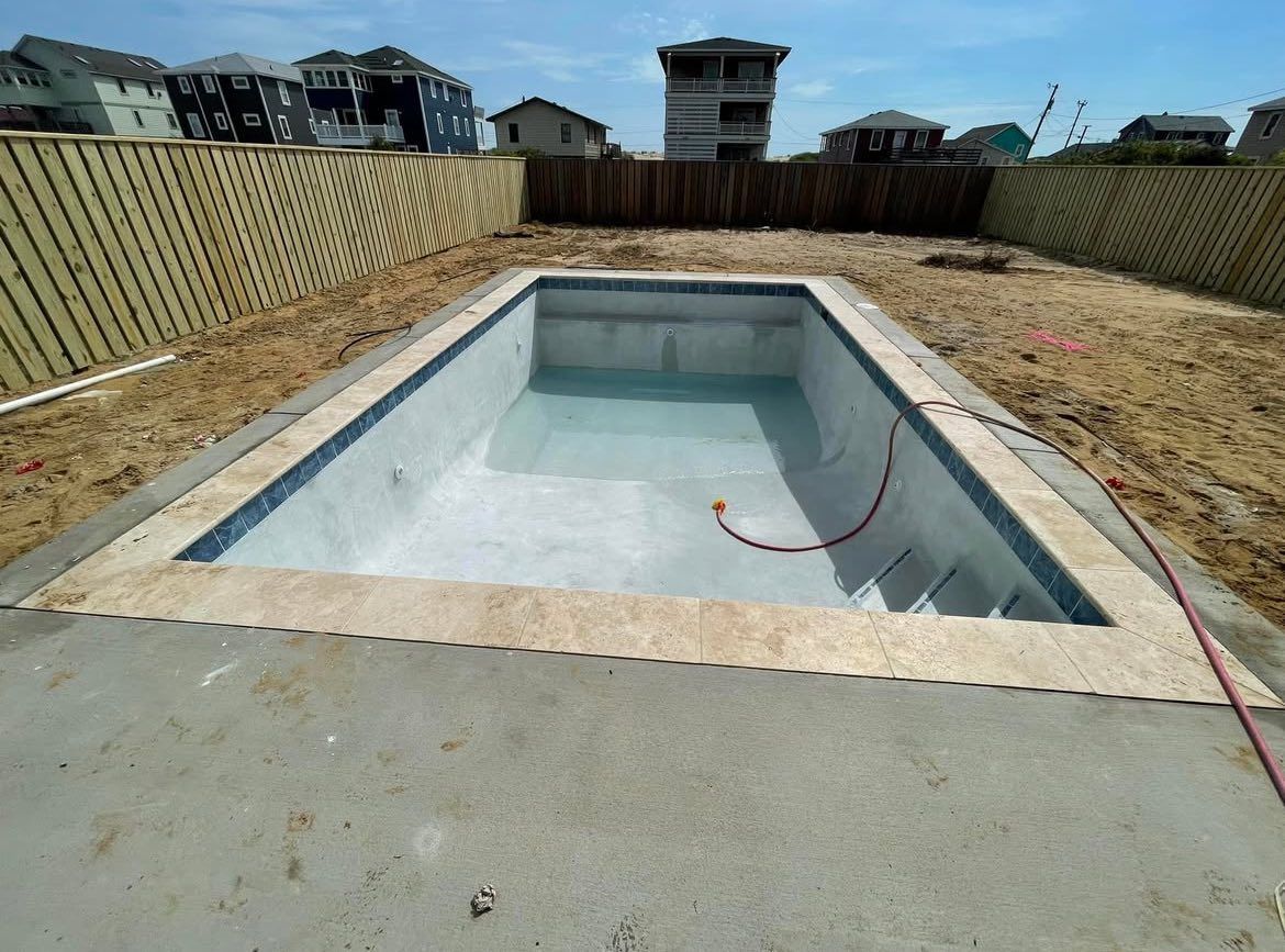 Empty rectangular swimming pool with tan coping and blue tile trim, surrounded by a fence and a dirt yard.