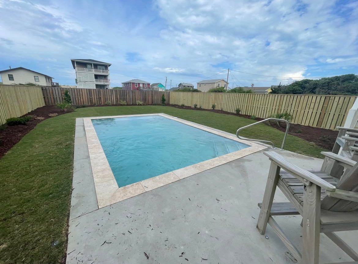 Rectangular pool in a backyard with green grass, a wooden fence, and blue sky.